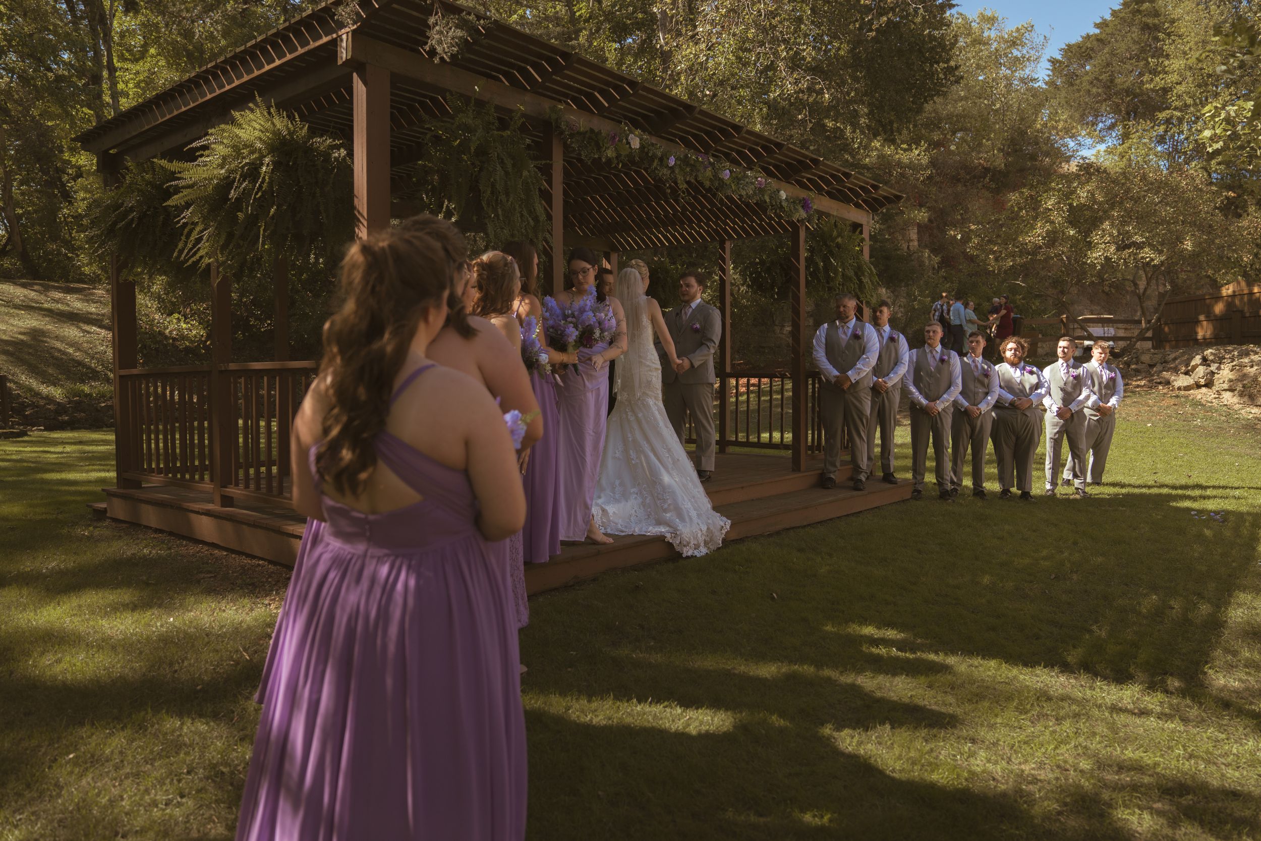 Wedding ceremony guests gathering under trees in outdoor garden setting.