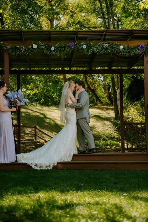 Wedding couple shares intimate moment under wooden pergola in lush garden setting.