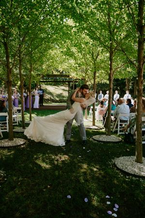 Outdoor wedding ceremony in tree-lined venue with guests seated on white chairs.