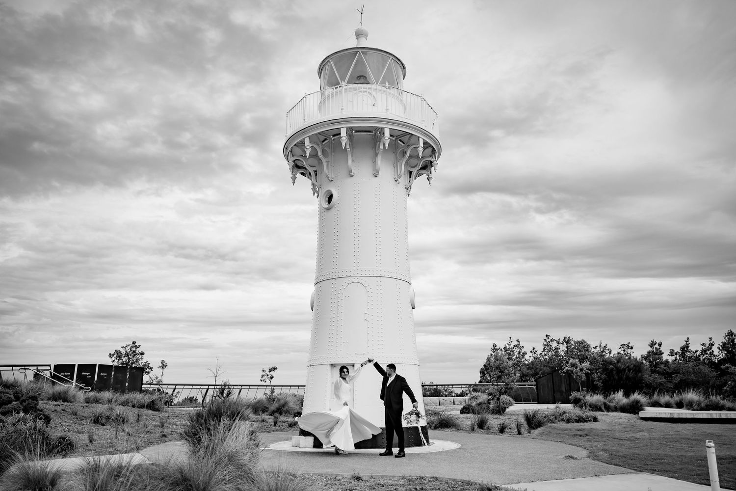 A white lighthouse stands tall against dramatic cloudy skies with silhouettes of visitors at its base.