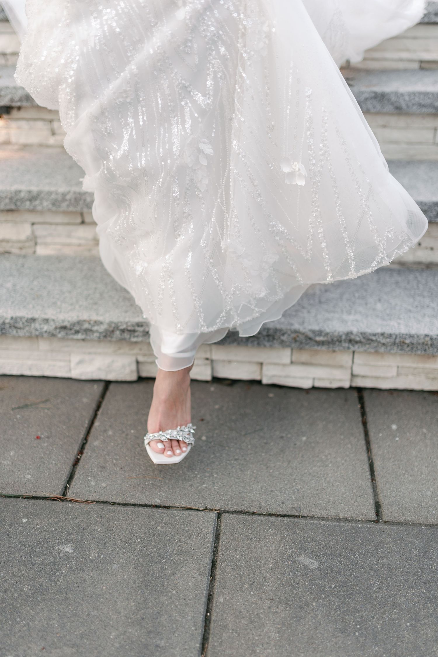 A detailed close-up of a bride's sparkly sandal peeking out beneath the hem of a flowing white wedding dress on stone steps.