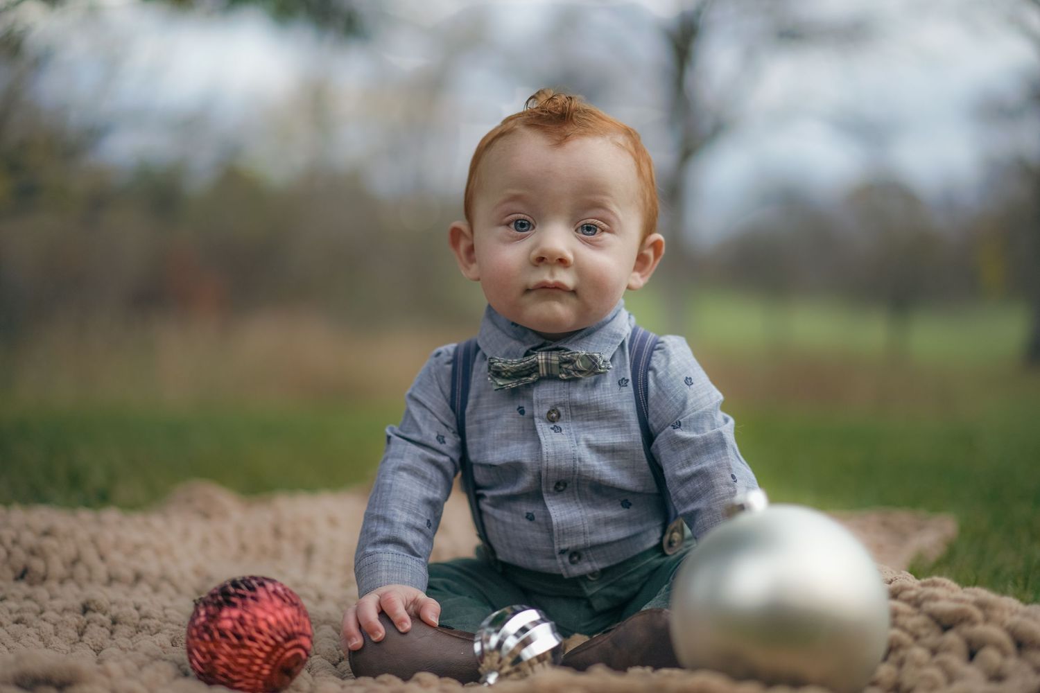 Baby dressed in bow tie and suspenders sits on ground with red and silver Christmas ornaments in outdoor setting. Michigan photographer.