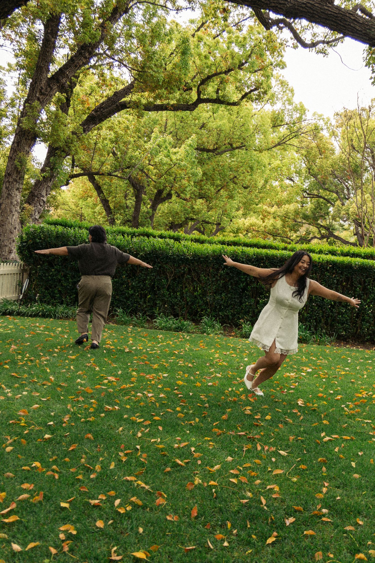 Two people playing and twirling in a lush garden with autumn leaves scattered on green grass.