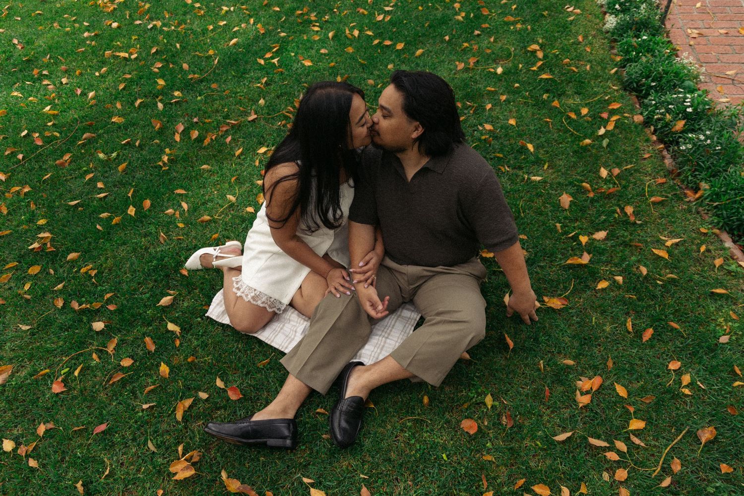 A couple shares a tender moment while sitting on grass covered with autumn leaves.