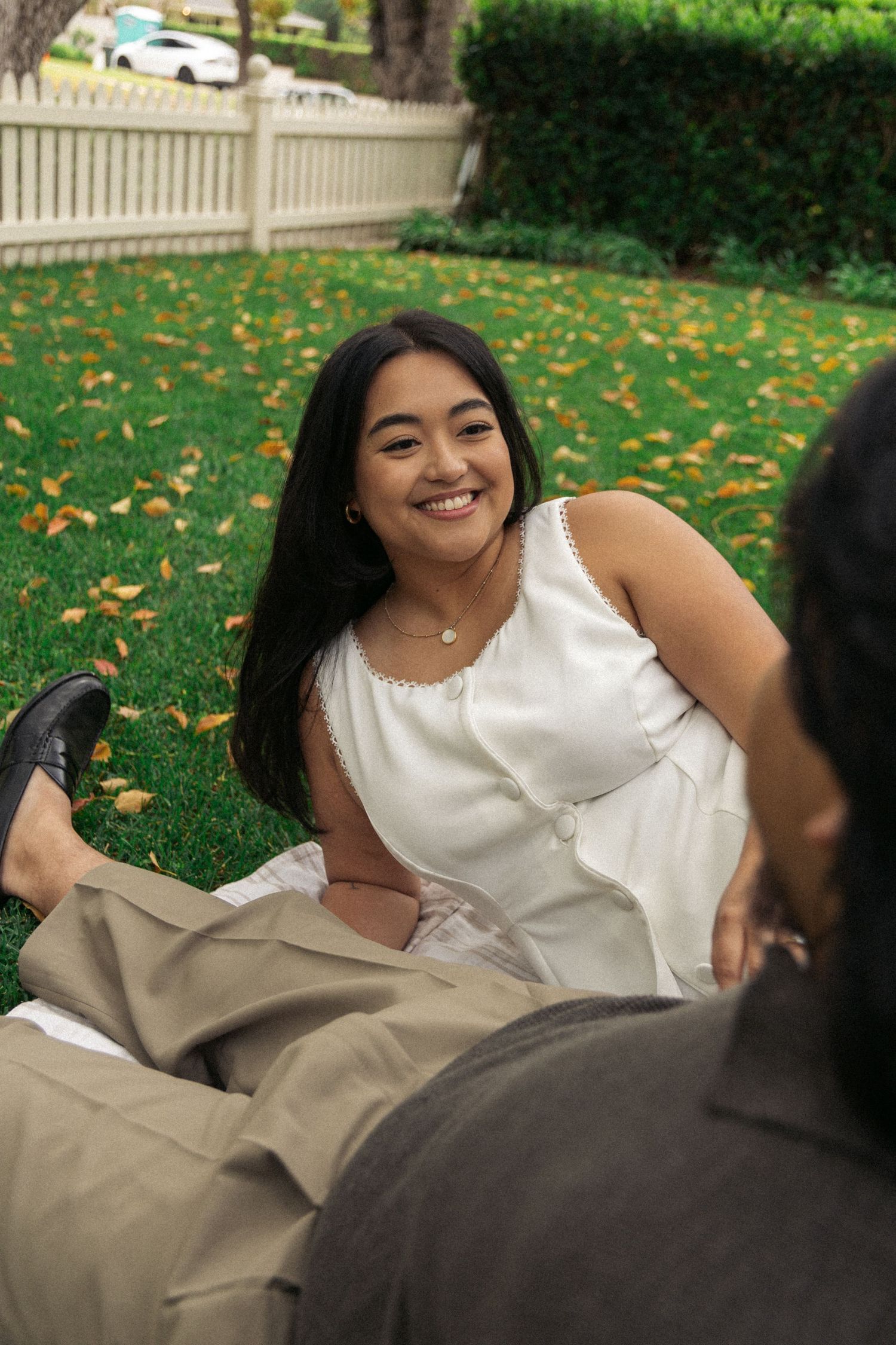 Person in white dress lying on grass during autumn with fallen leaves and white fence in background.