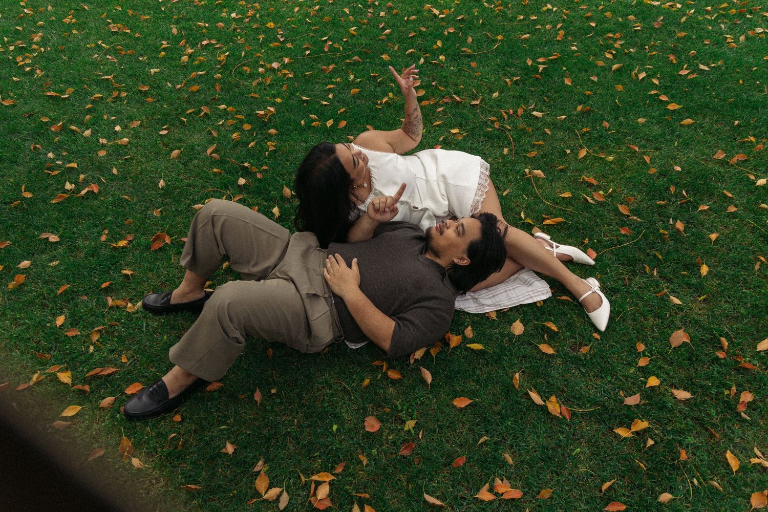 People lying in fallen leaves on grass beneath trees on an autumn day.
