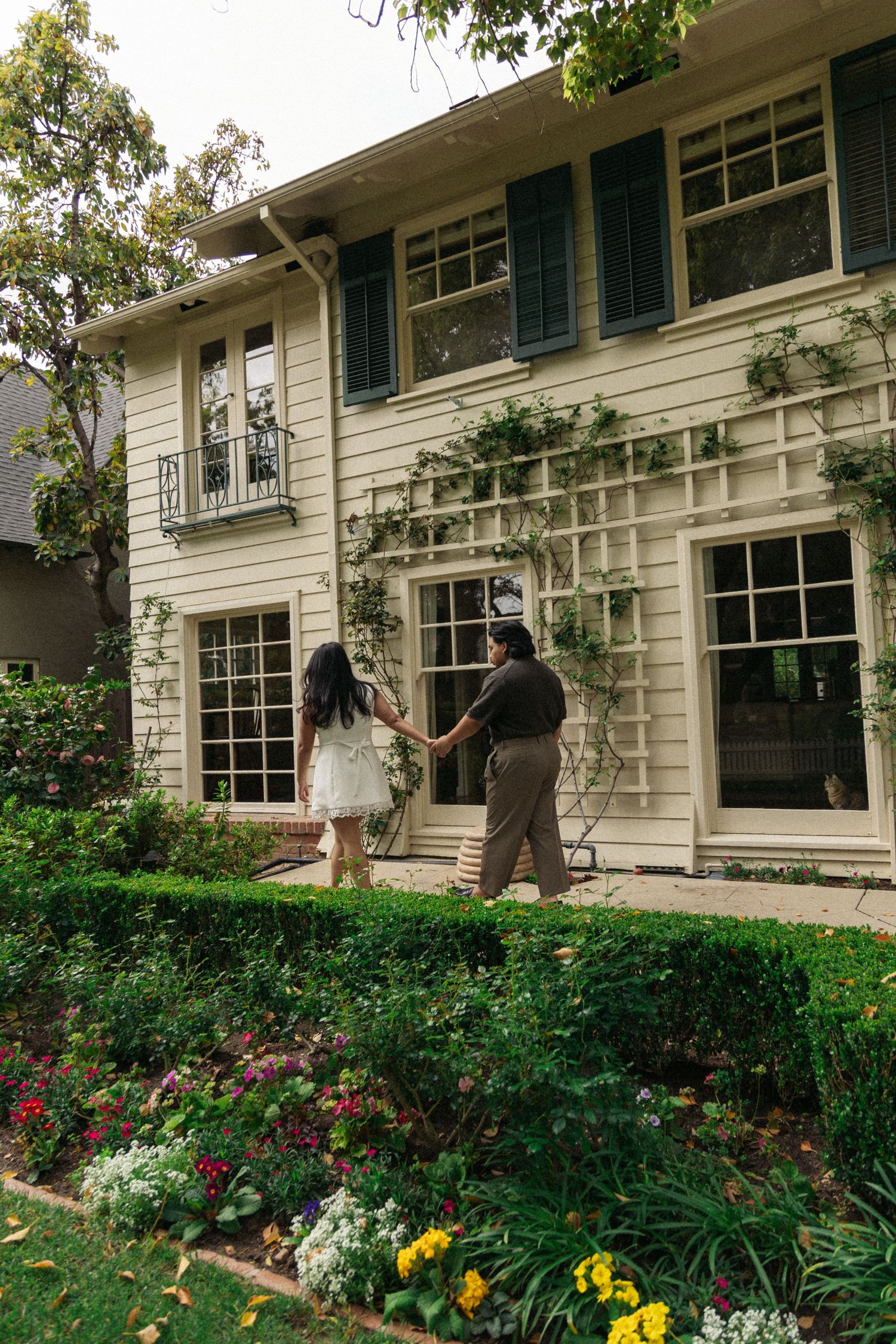 Two figures standing in front of a white colonial house with flowering garden beds.