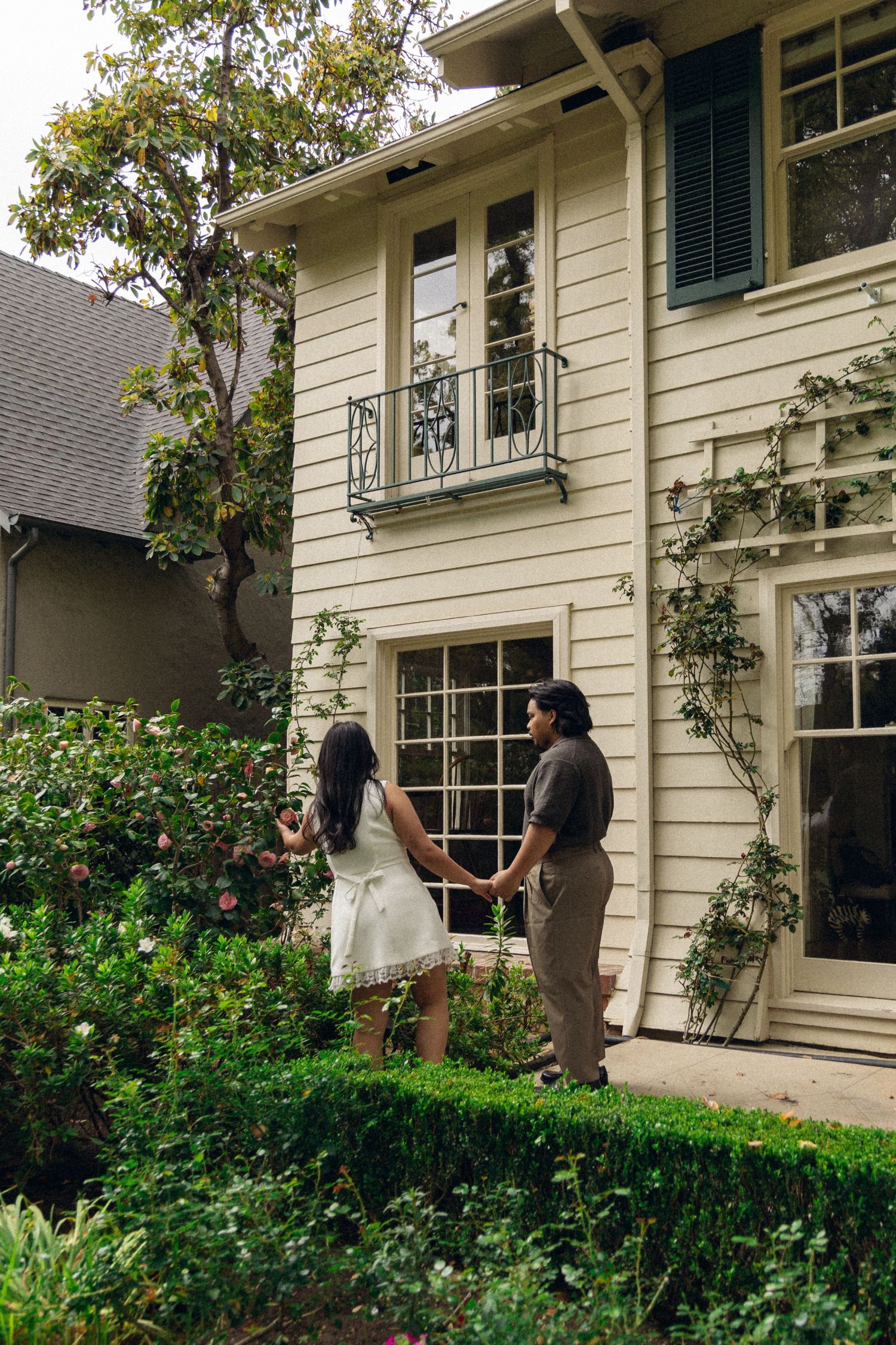 A couple holding hands while admiring a charming white colonial-style house with green shutters and climbing vines.