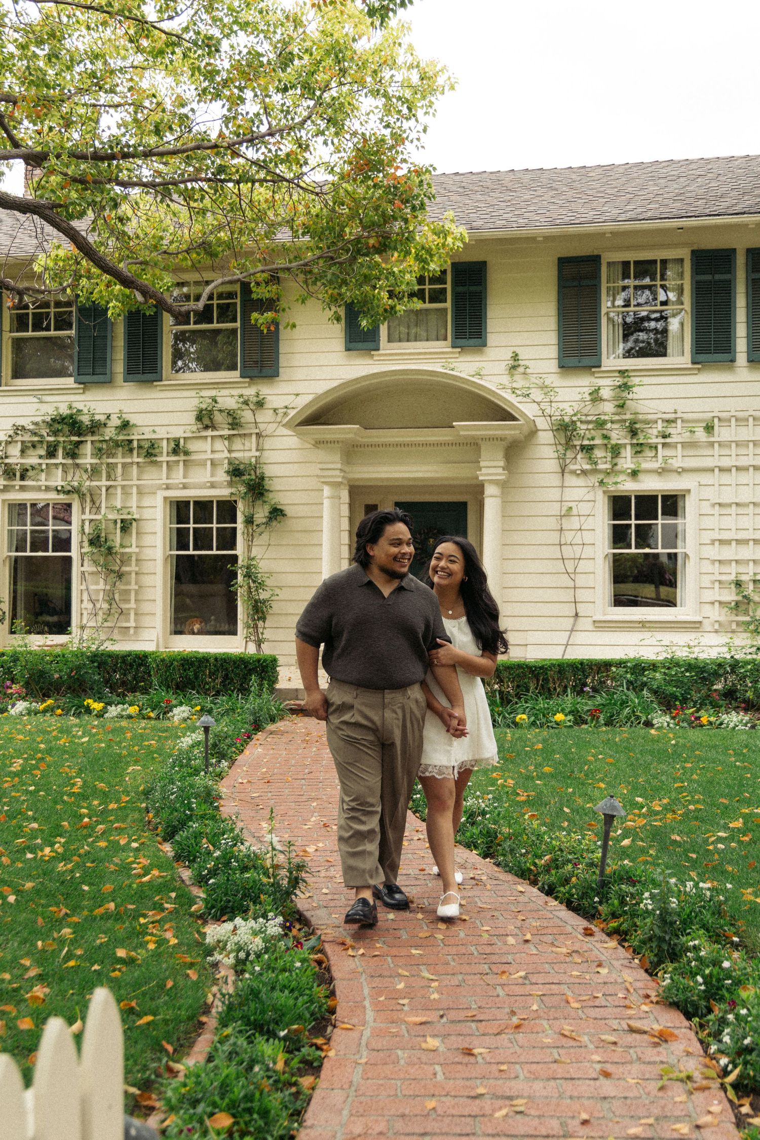 A romantic couple stands on a brick pathway in front of a classic white colonial house.