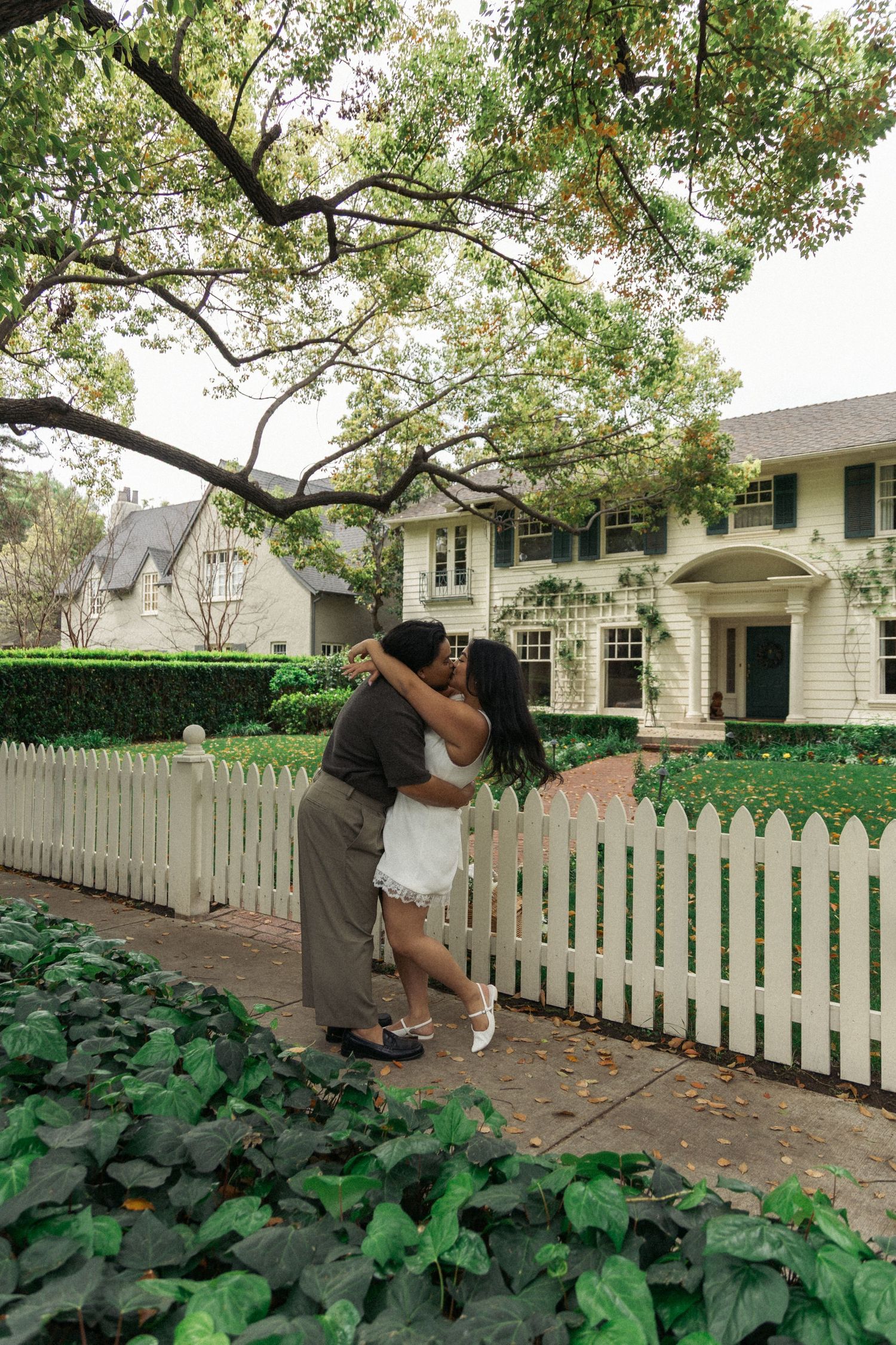 A romantic moment shared in front of a classic white colonial house with a picket fence.