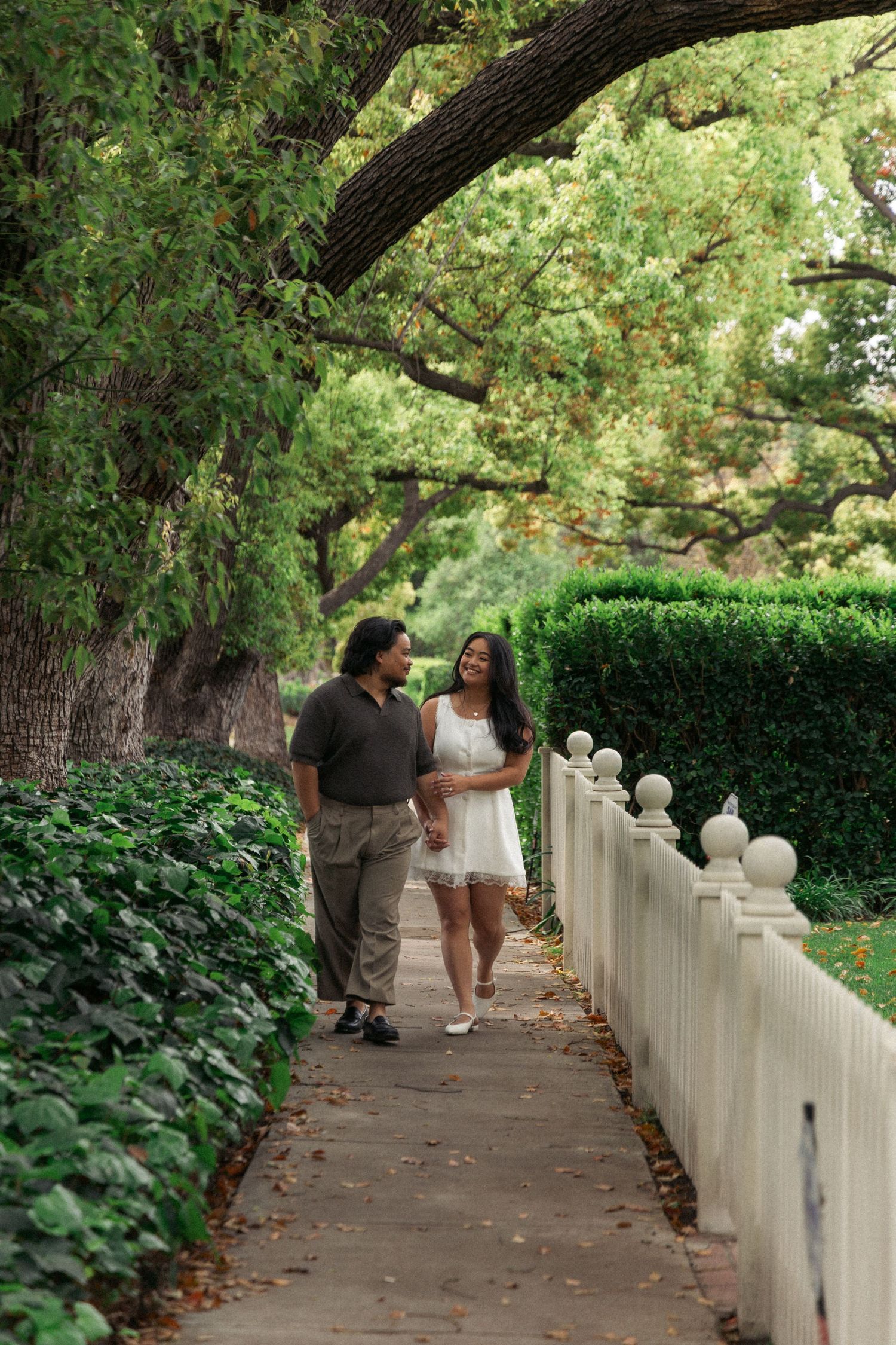A couple strolls along a picturesque garden path with a white picket fence and overhanging oak trees.