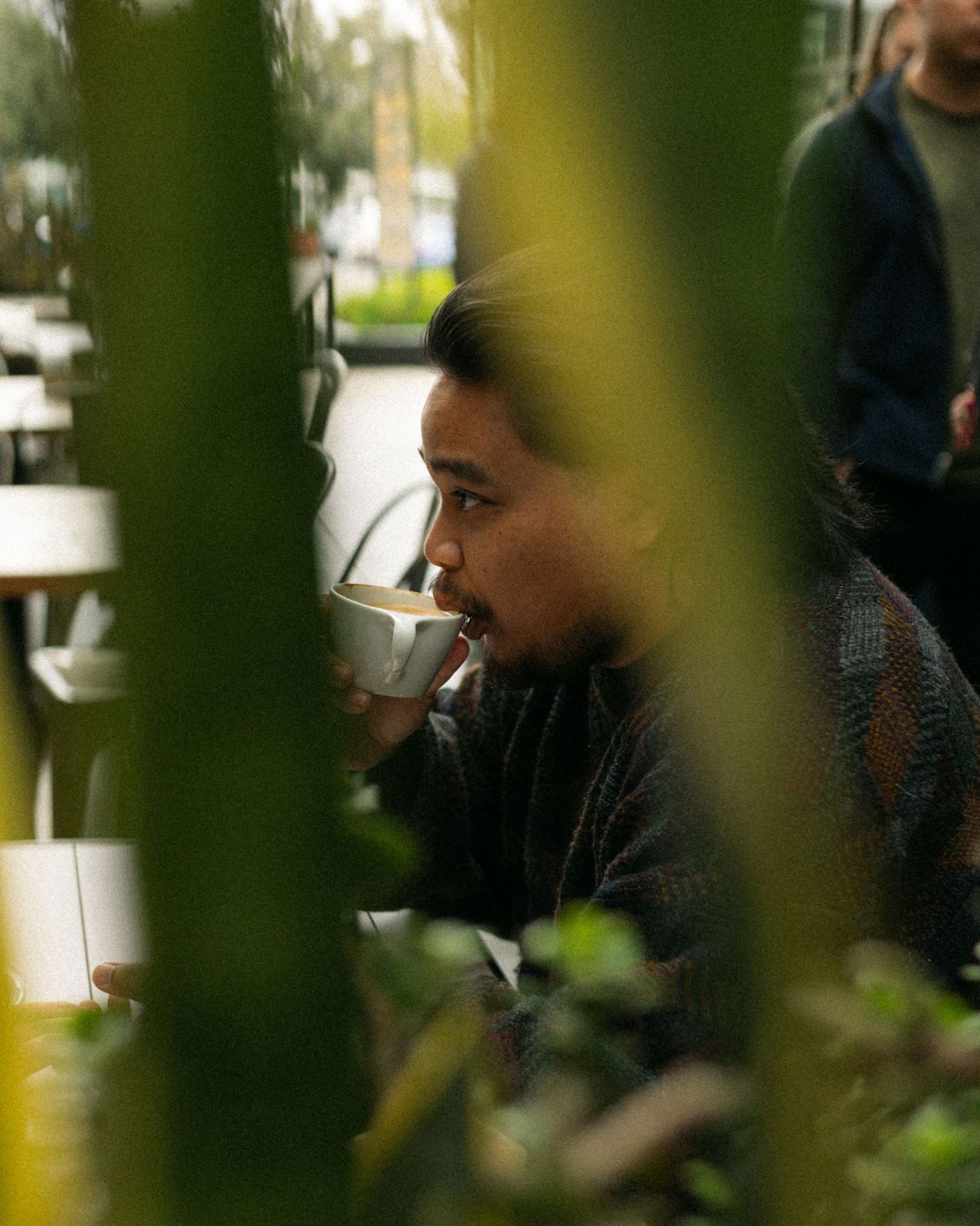 A candid shot through foliage shows someone enjoying a drink at an outdoor cafe.