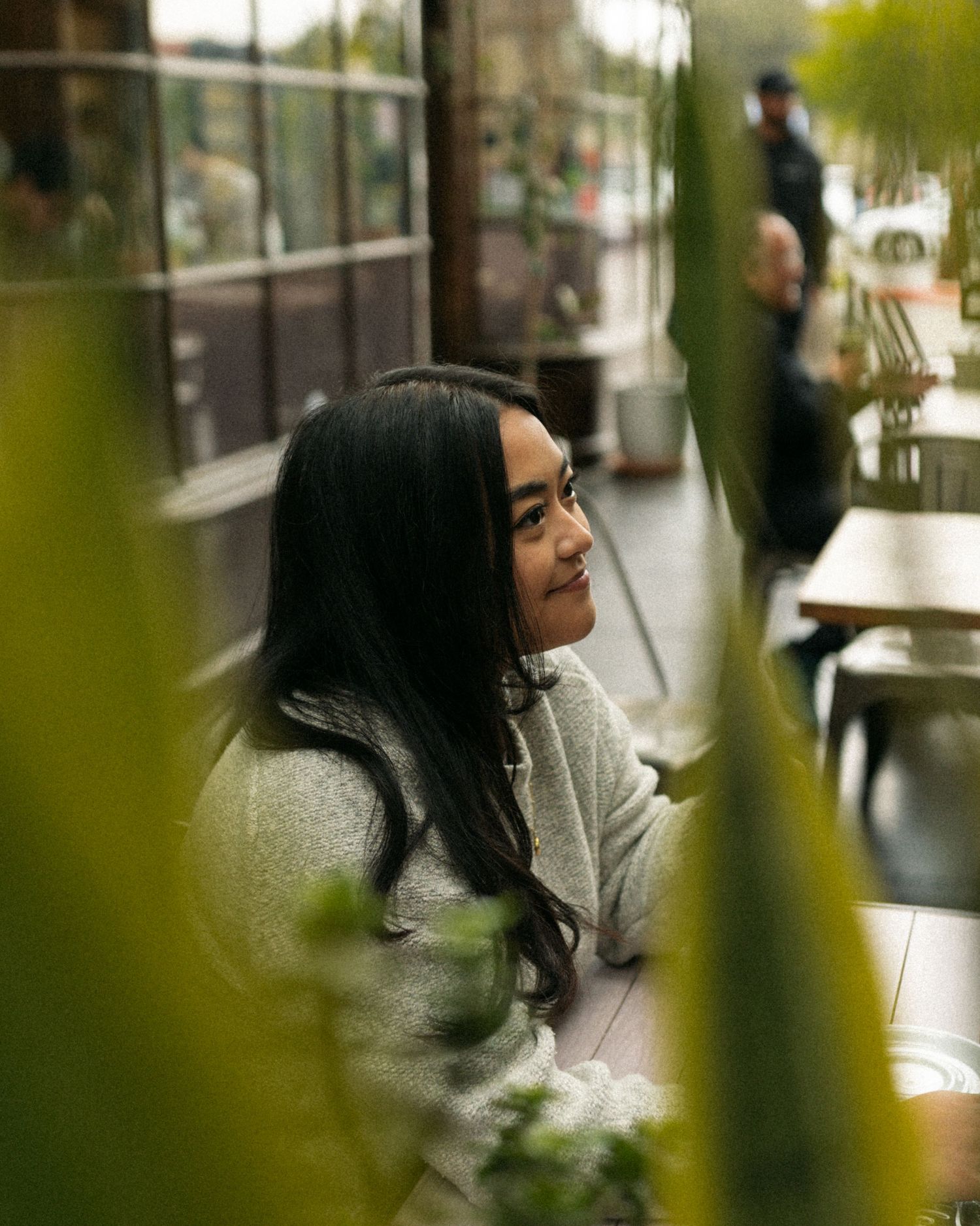A person in a gray sweater sits thoughtfully at a cafe table seen through green foliage.