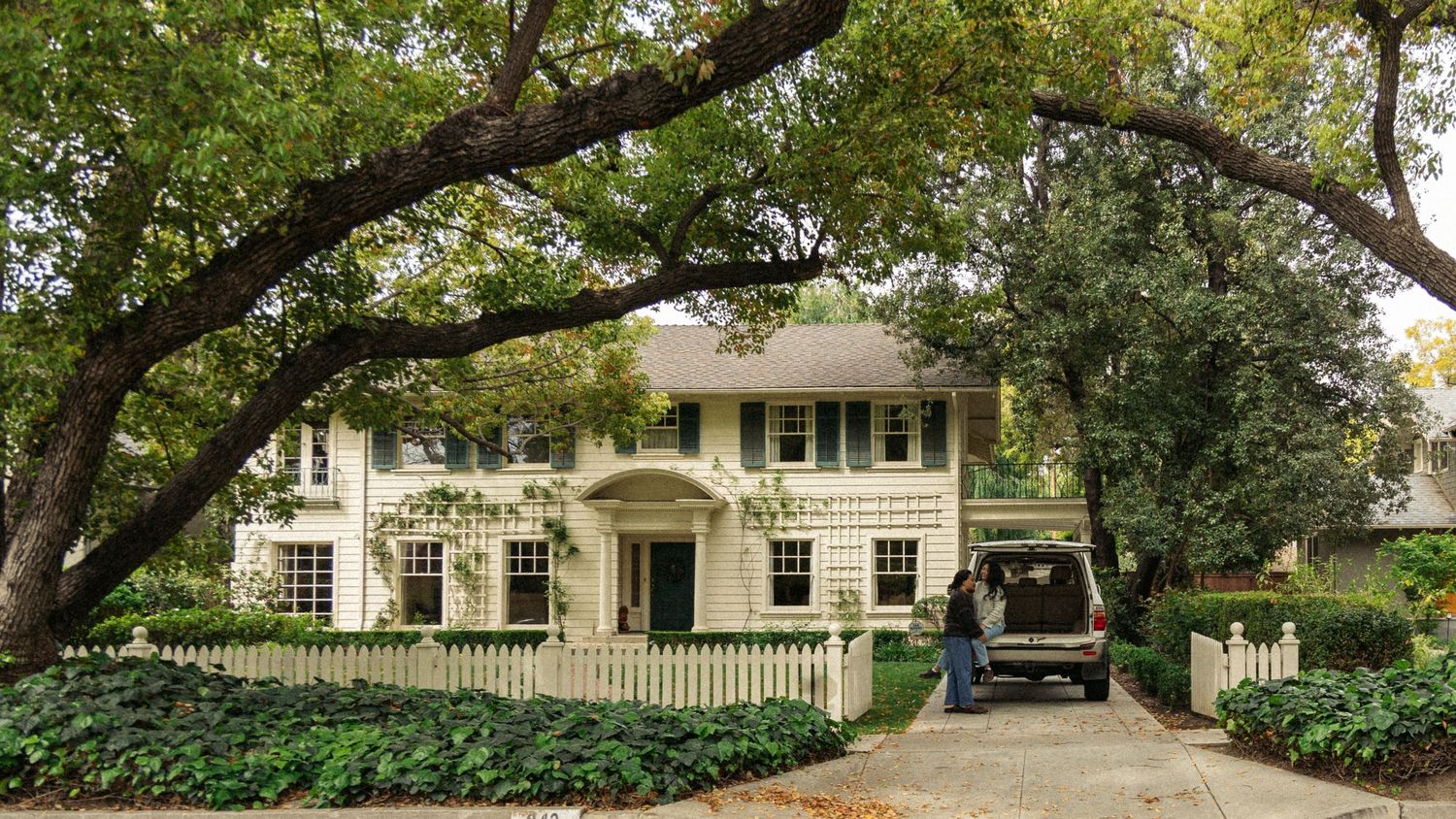 Beautiful colonial-style white house with green shutters and white picket fence surrounded by lush landscaping.