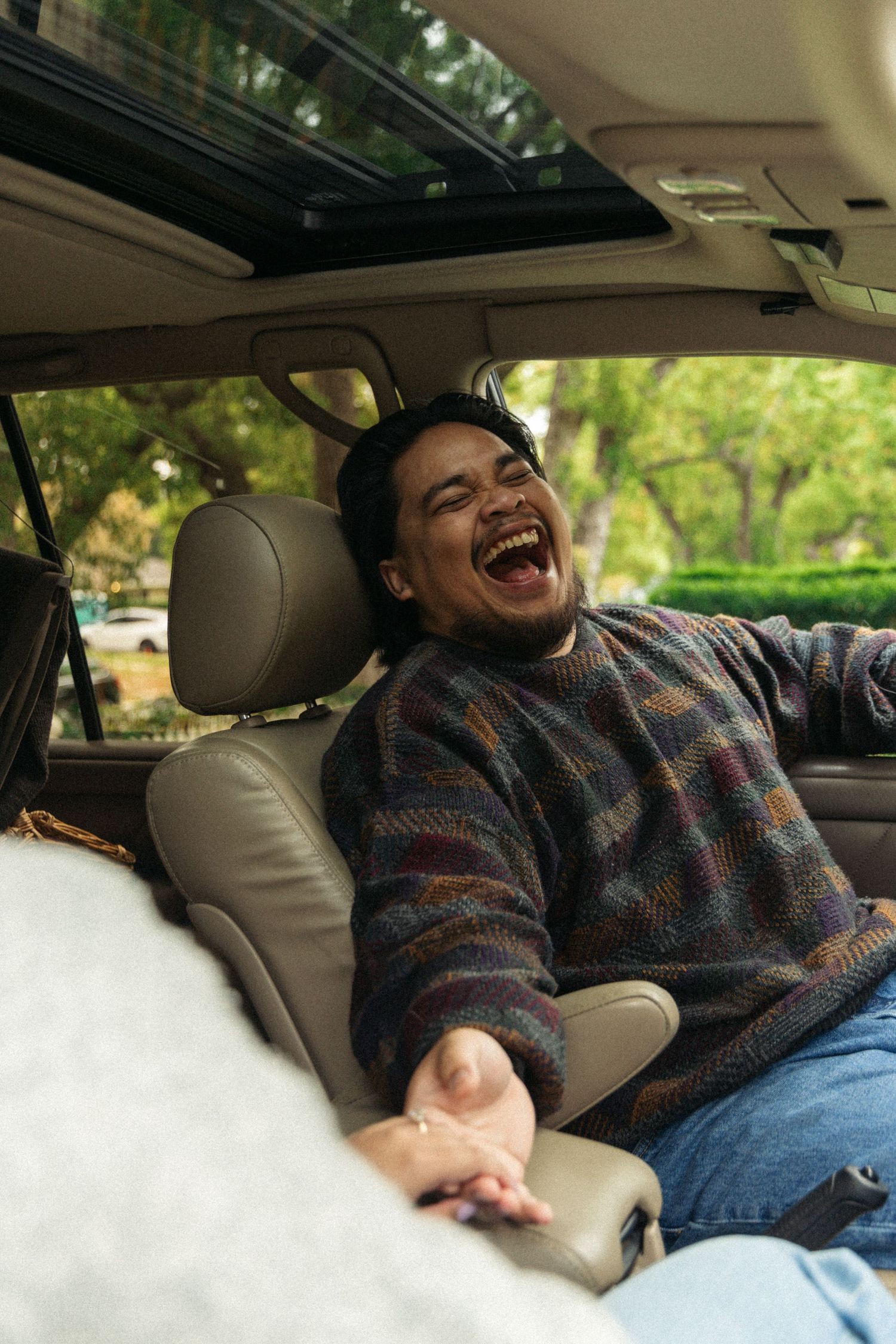 Person laughing joyfully while relaxing in car backseat wearing patterned sweater and jeans.