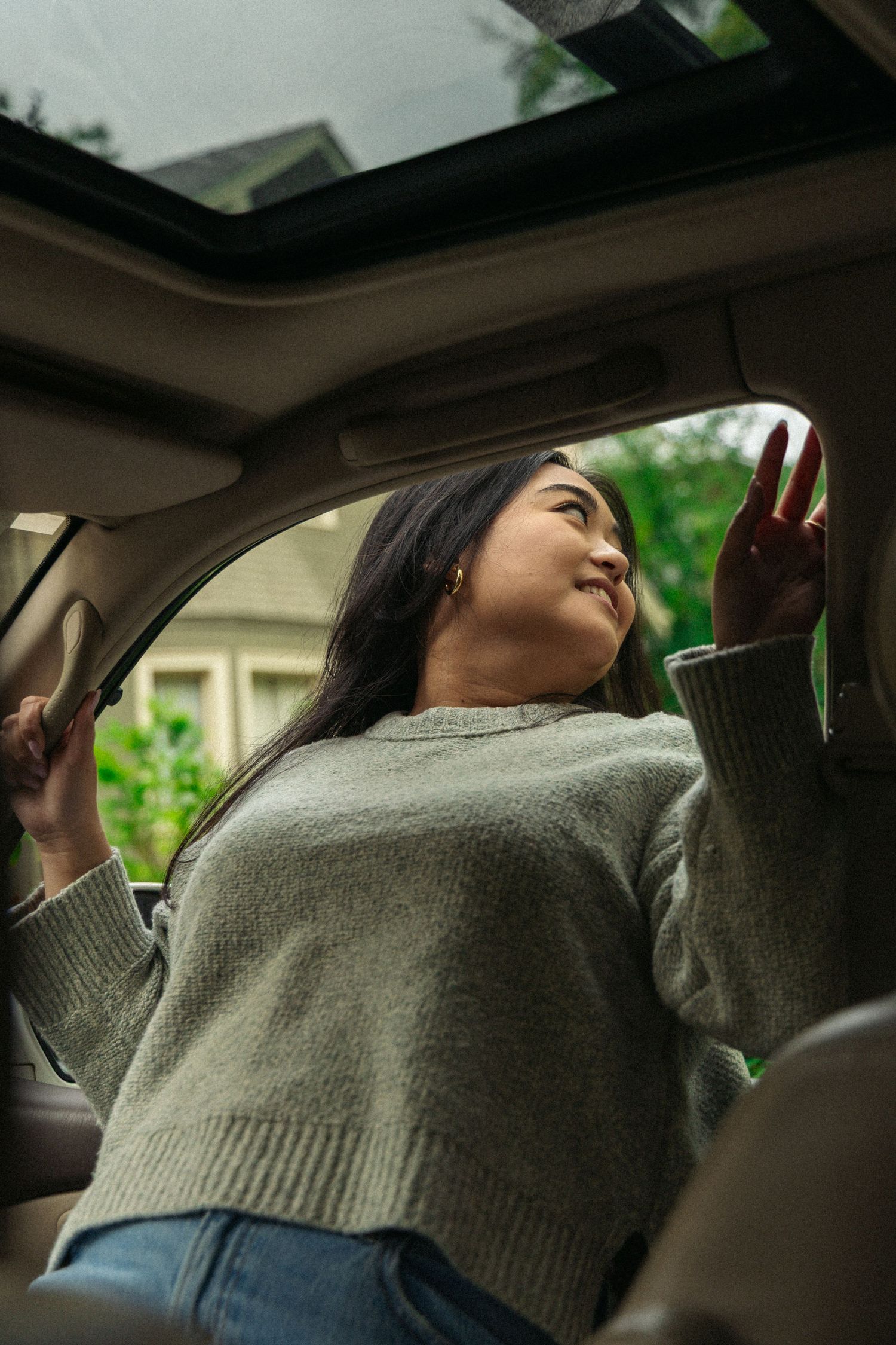 Person in grey sweater reaching up through car sunroof on sunny day.