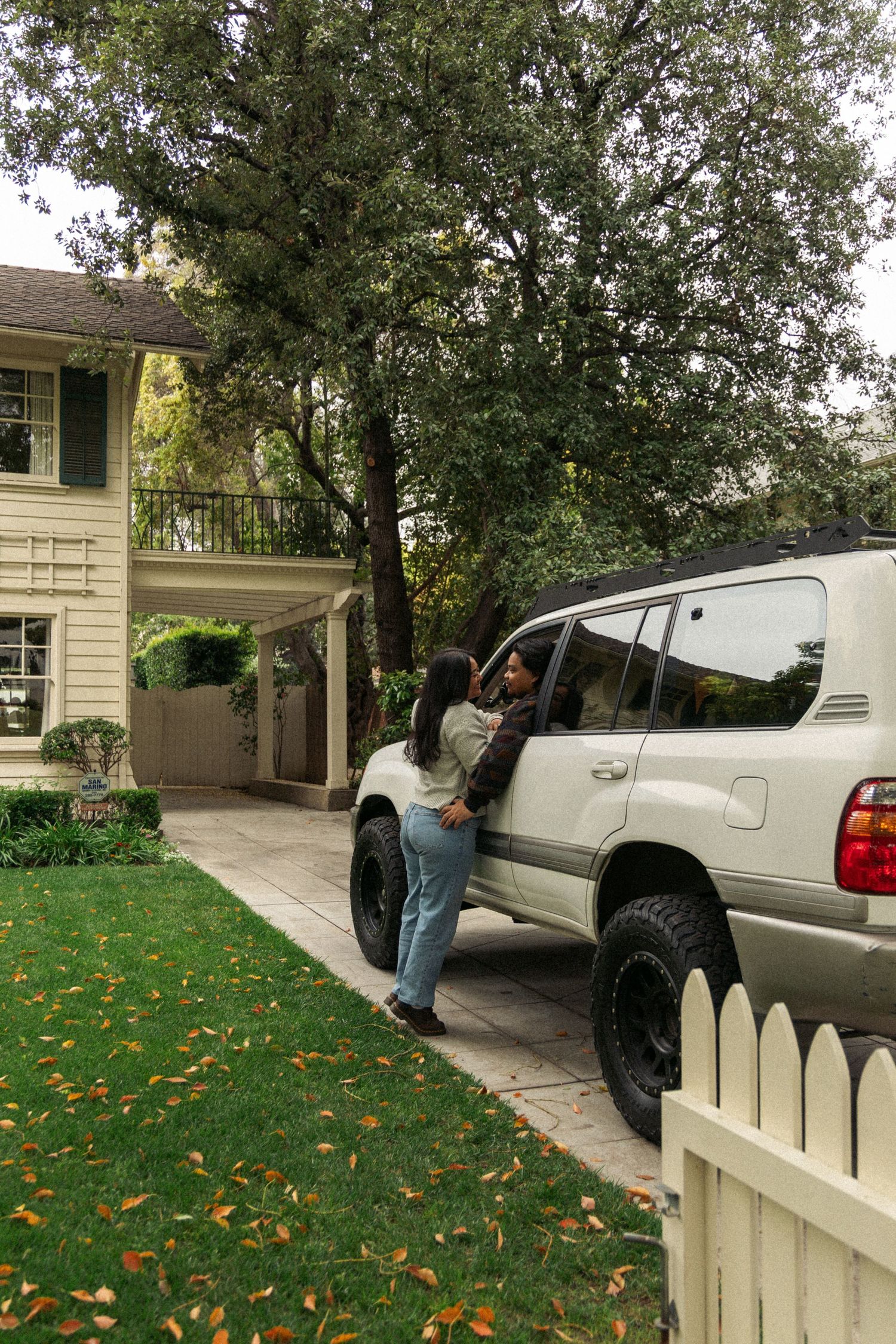 People interact near an SUV parked in front of a beautiful suburban home with fall foliage.