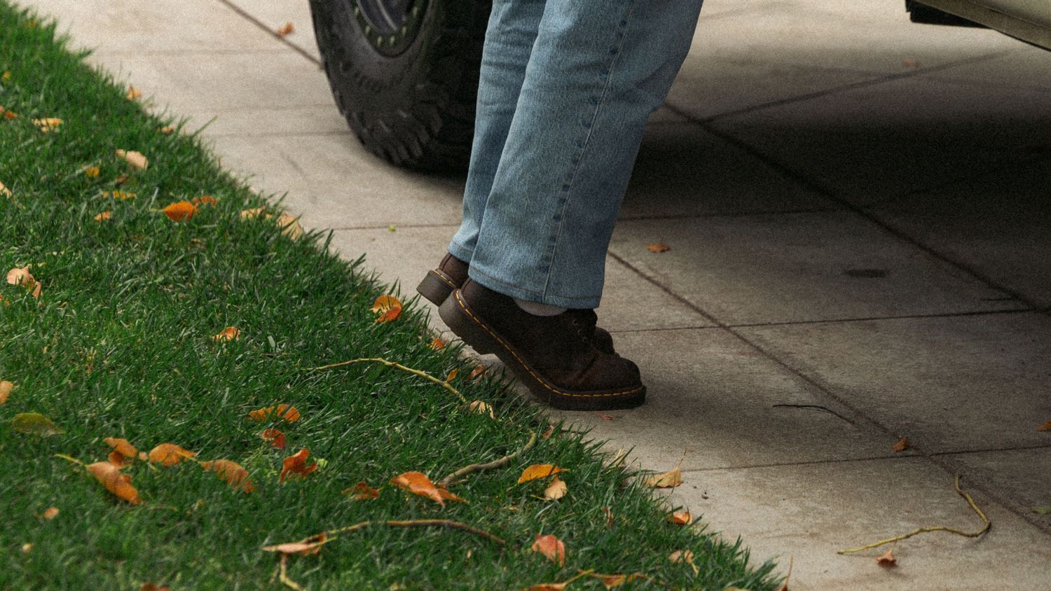 Brown shoes walking on sidewalk next to grass with scattered fall leaves.