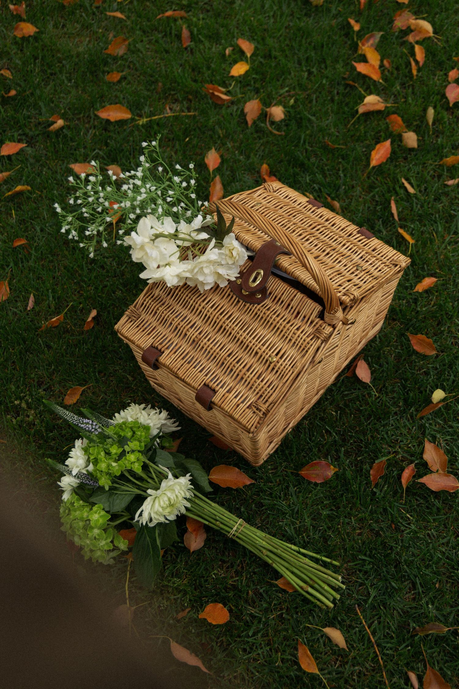 Wicker picnic basket with white flowers and scattered autumn leaves on green grass.