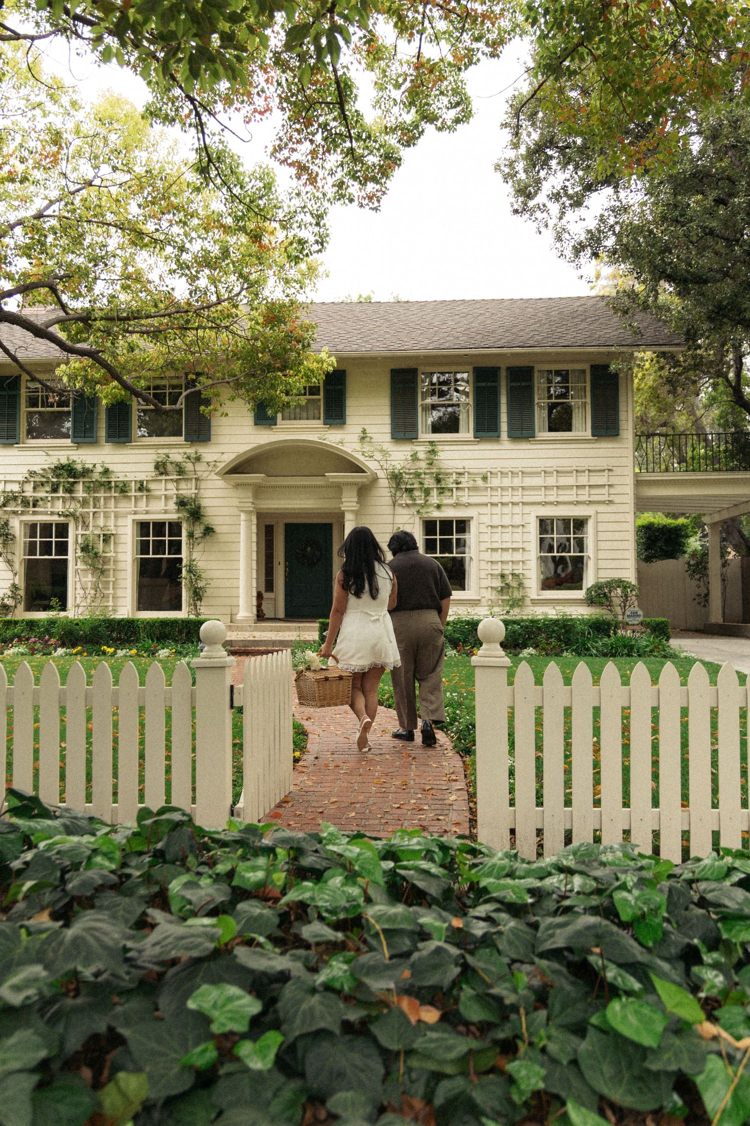 A couple walks toward a white colonial house with green shutters and a picket fence.