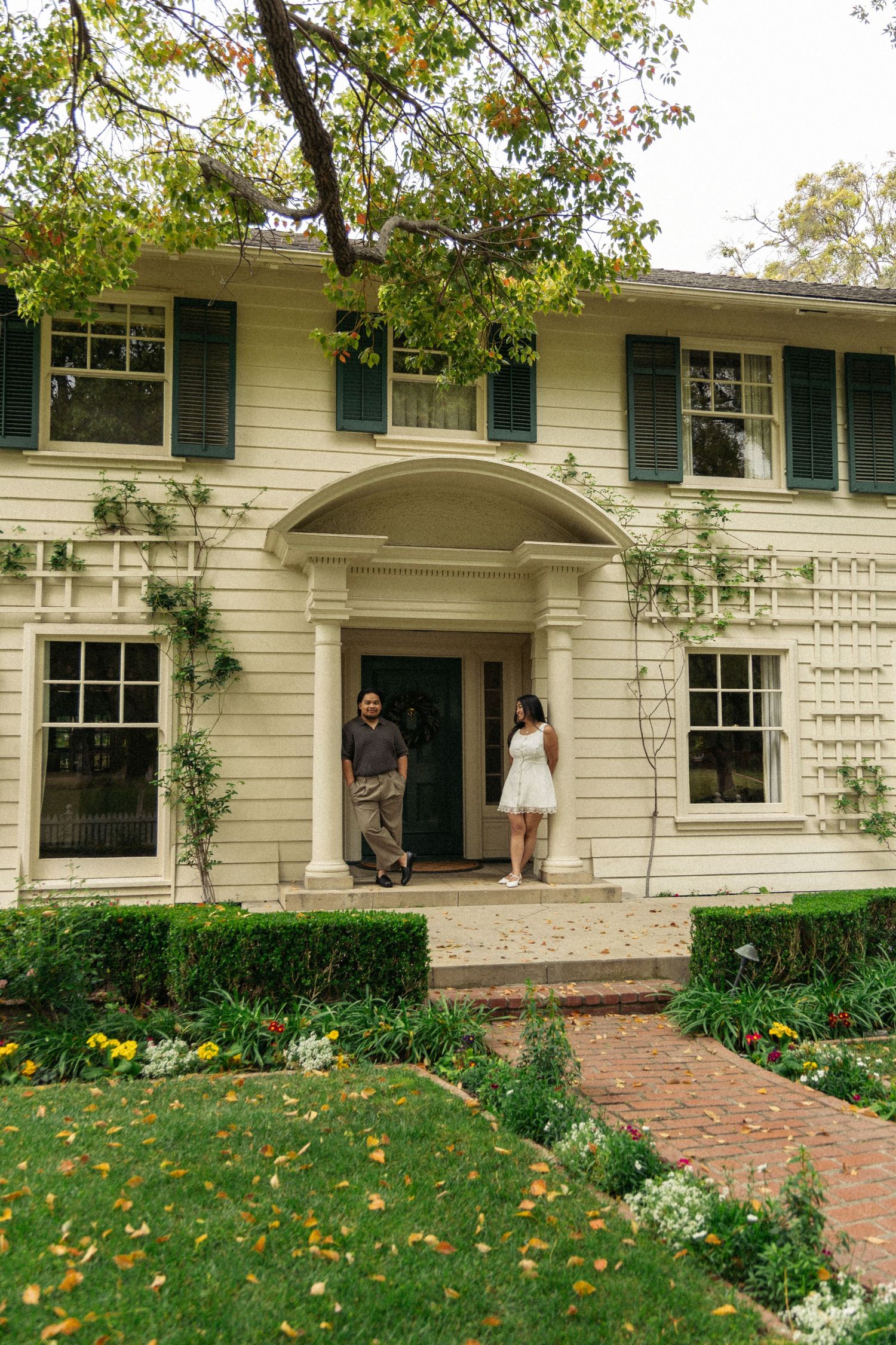 White colonial style house with green shutters and brick walkway surrounded by gardens.