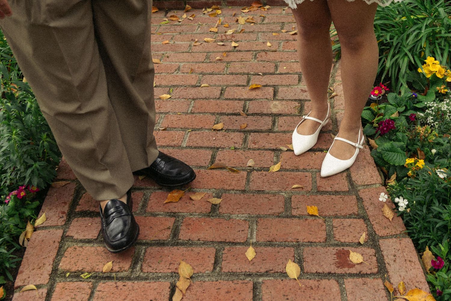 Contrasting formal and casual shoes on red brick pathway with garden flowers.