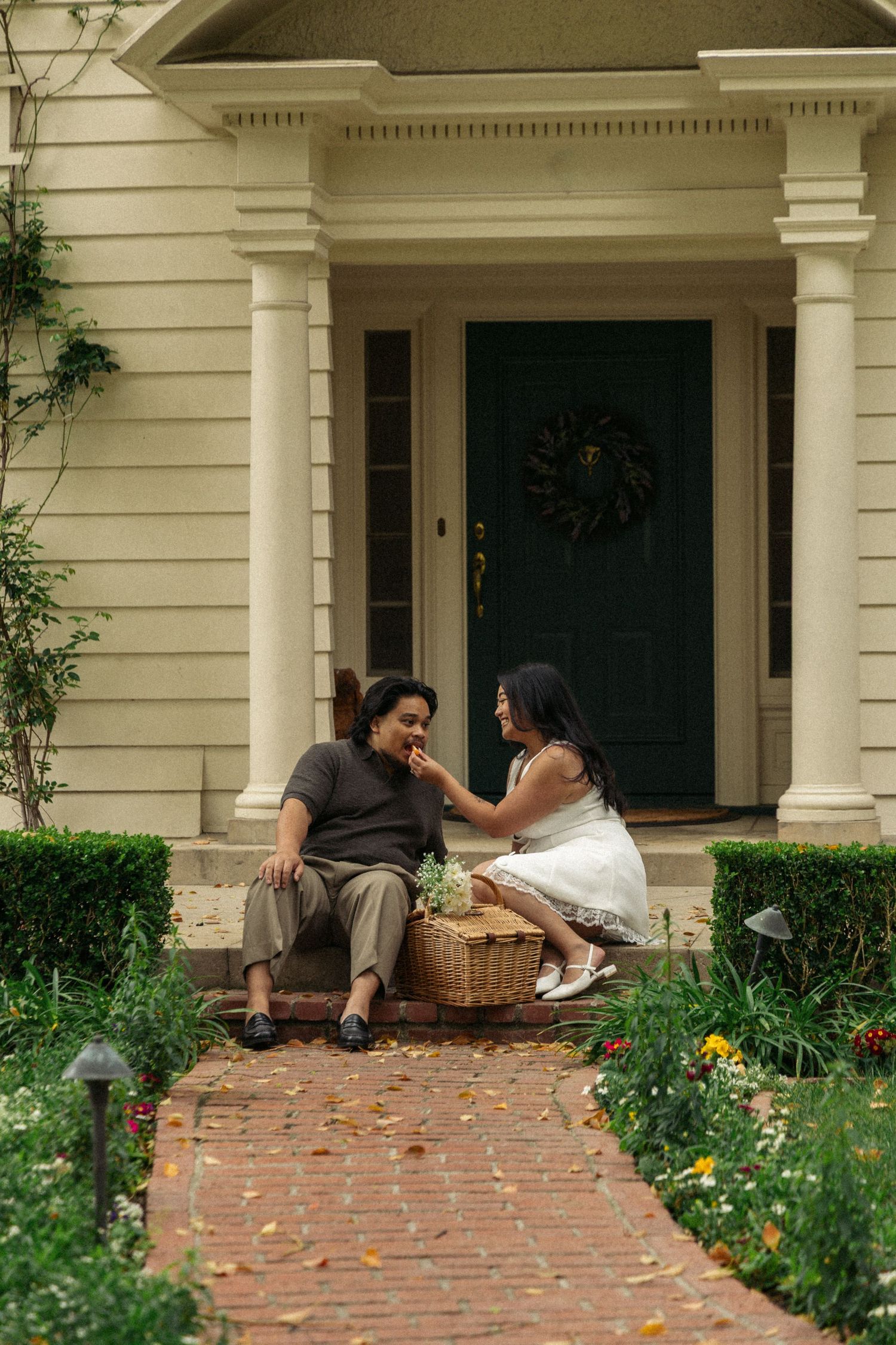 Two people share a picnic on brick steps of a classic white colonial home with black door and wreath.