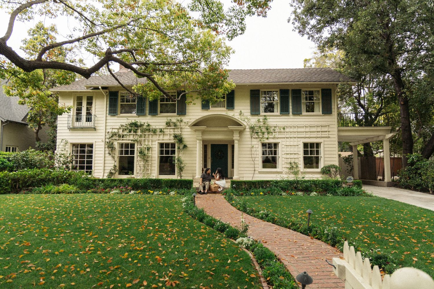 Classic white two-story colonial house with symmetrical windows and brick pathway through autumn leaves.