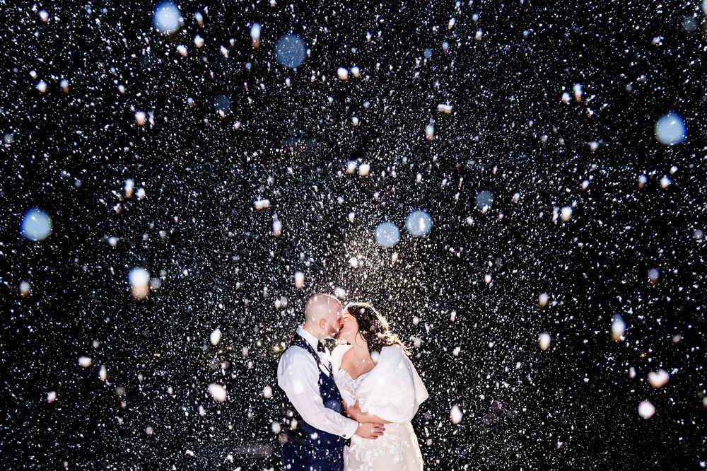 A romantic couple embraces under falling snow at night creating a magical winter wedding moment.