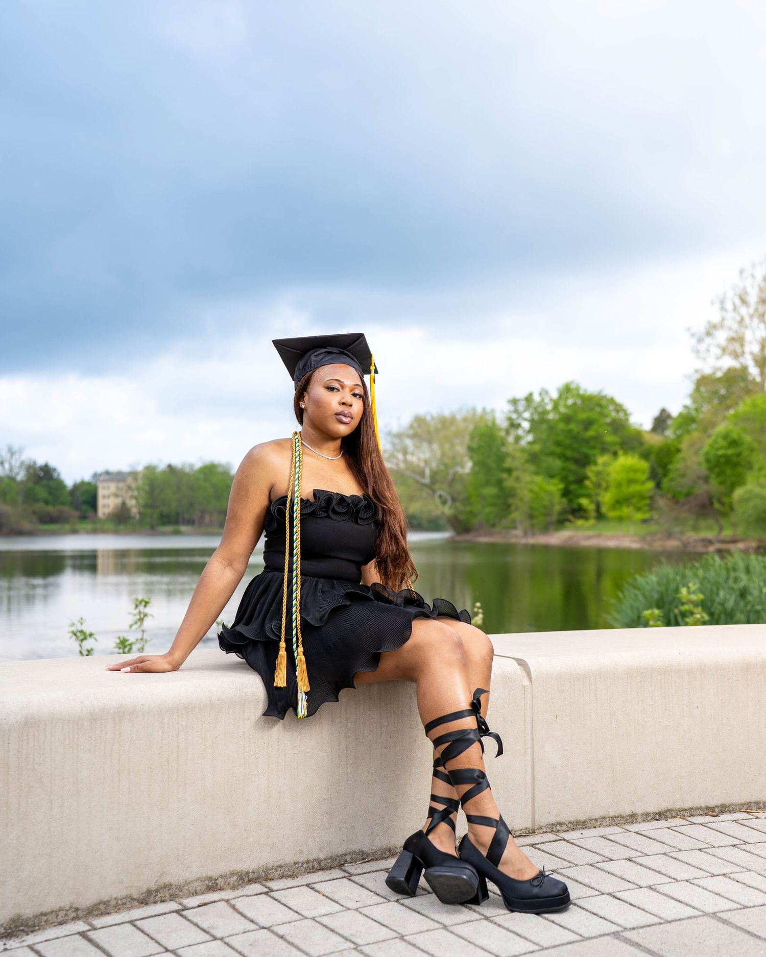Graduate poses in black dress and platform gladiator sandals by lake on graduation day.