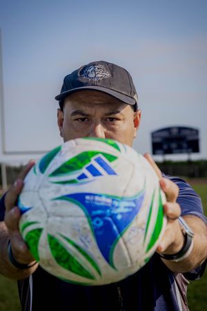Close-up shot of a rugby ball with blue and green stripes being held up against a blue sky background.