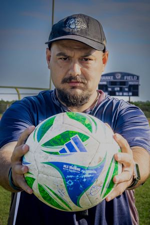 Soccer coach holds blue and green soccer ball while standing outdoors on athletic field with stadium backdrop.