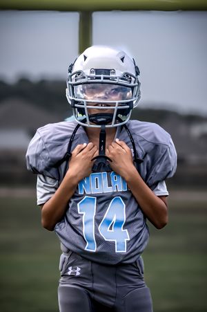 Youth football player wearing a gray Under Armour uniform with number 14 and white helmet stands on field.