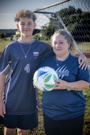 A soccer player wearing jersey number 4 stands next to their coach holding a white and green ball by a goal net.