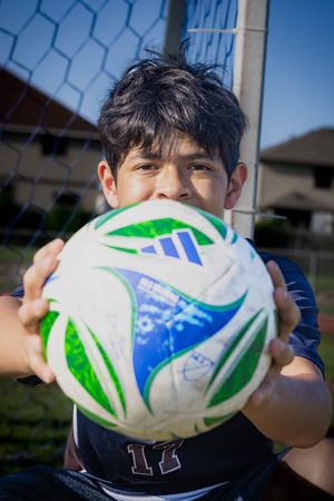 A soccer player holds up a white, green and blue soccer ball against a chain link fence background during practice.