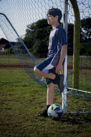 Soccer player stands by goal post with ball at sunset on grassy field wearing blue and white athletic gear.