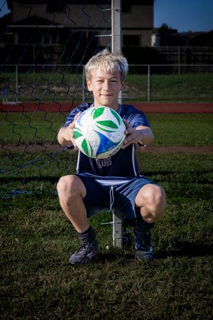 Young soccer player in blue uniform poses with green and white soccer ball in front of goal net on sports field.