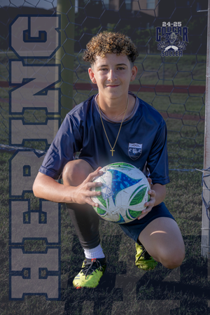 Soccer player in navy blue uniform kneels while holding a soccer ball on field with fluorescent yellow cleats.