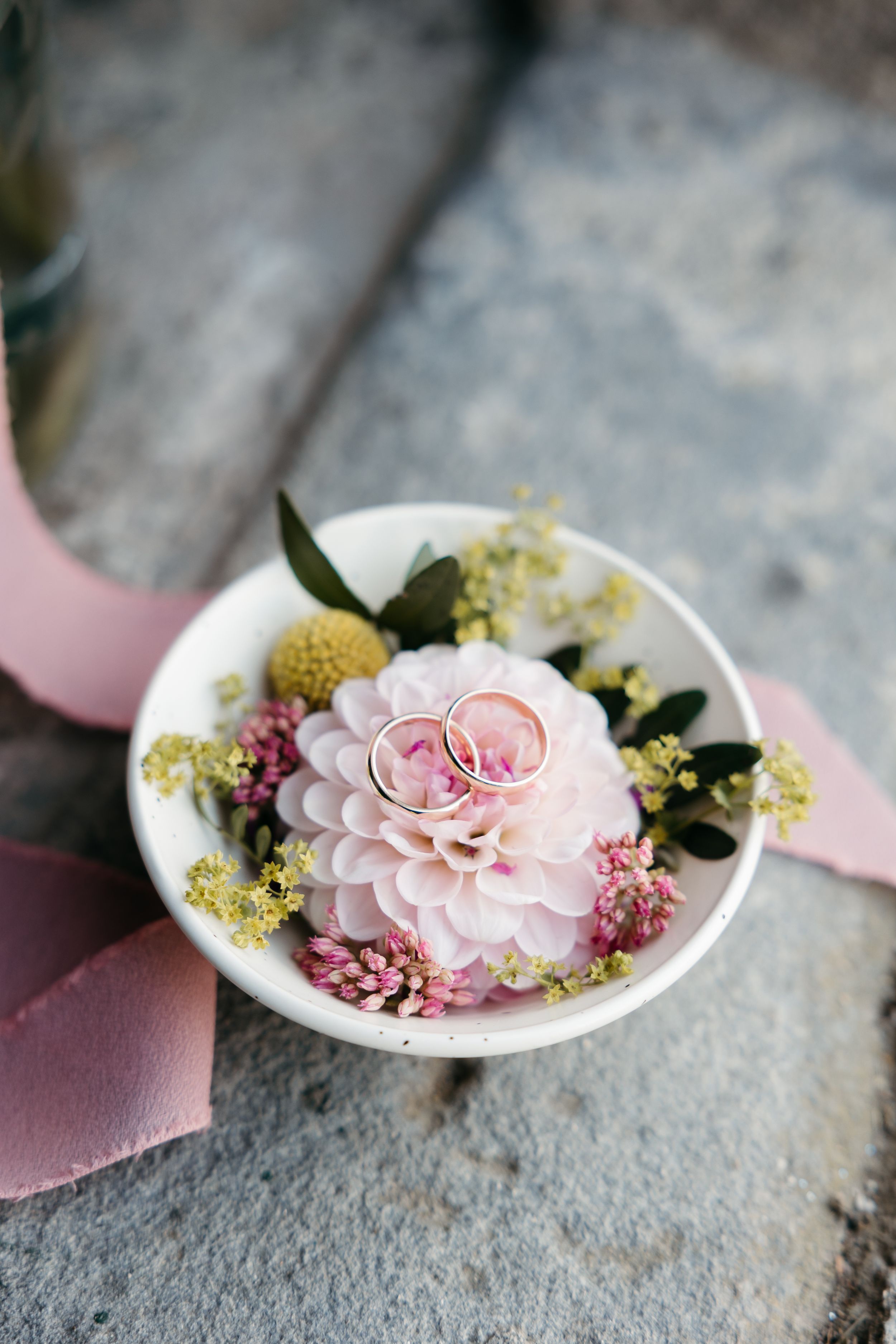 Wedding rings rest on a delicate pink dahlia flower surrounded by greenery and smaller blooms in a white dish.
