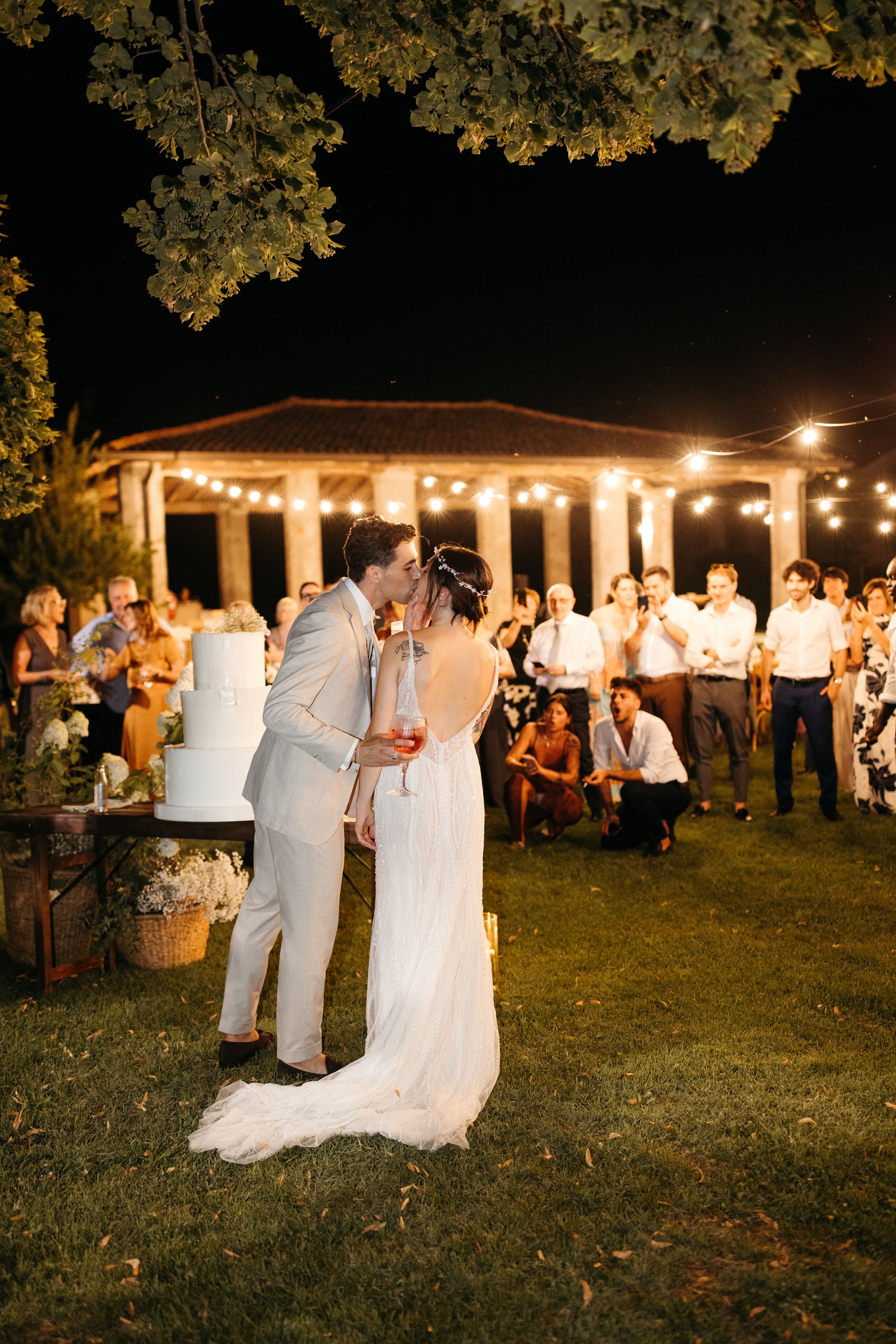 A couple shares a romantic moment at their evening outdoor wedding reception under a lit gazebo with guests in background.