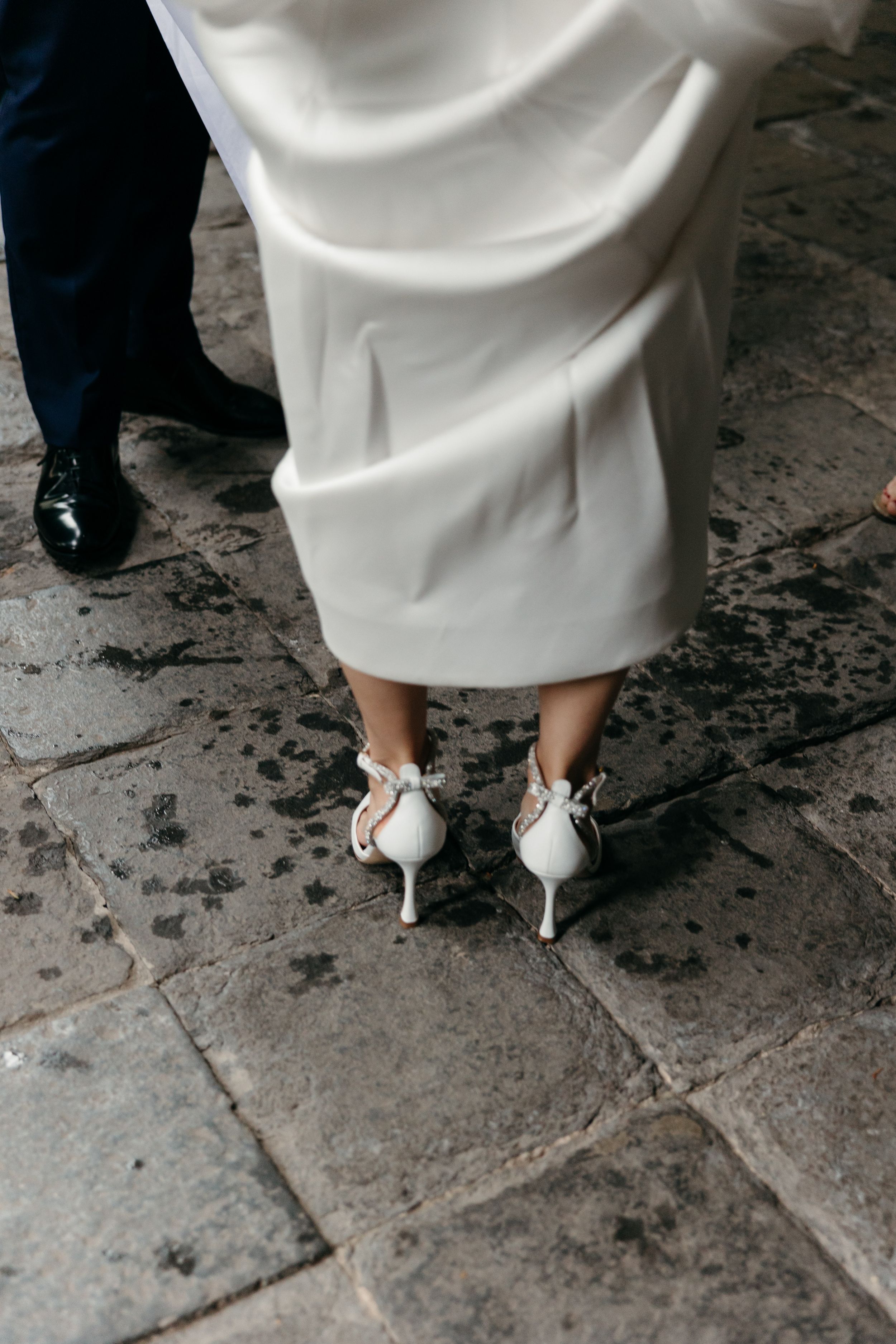 Close-up view of a bride wearing elegant white heels and a flowing wedding dress on stone pavement.