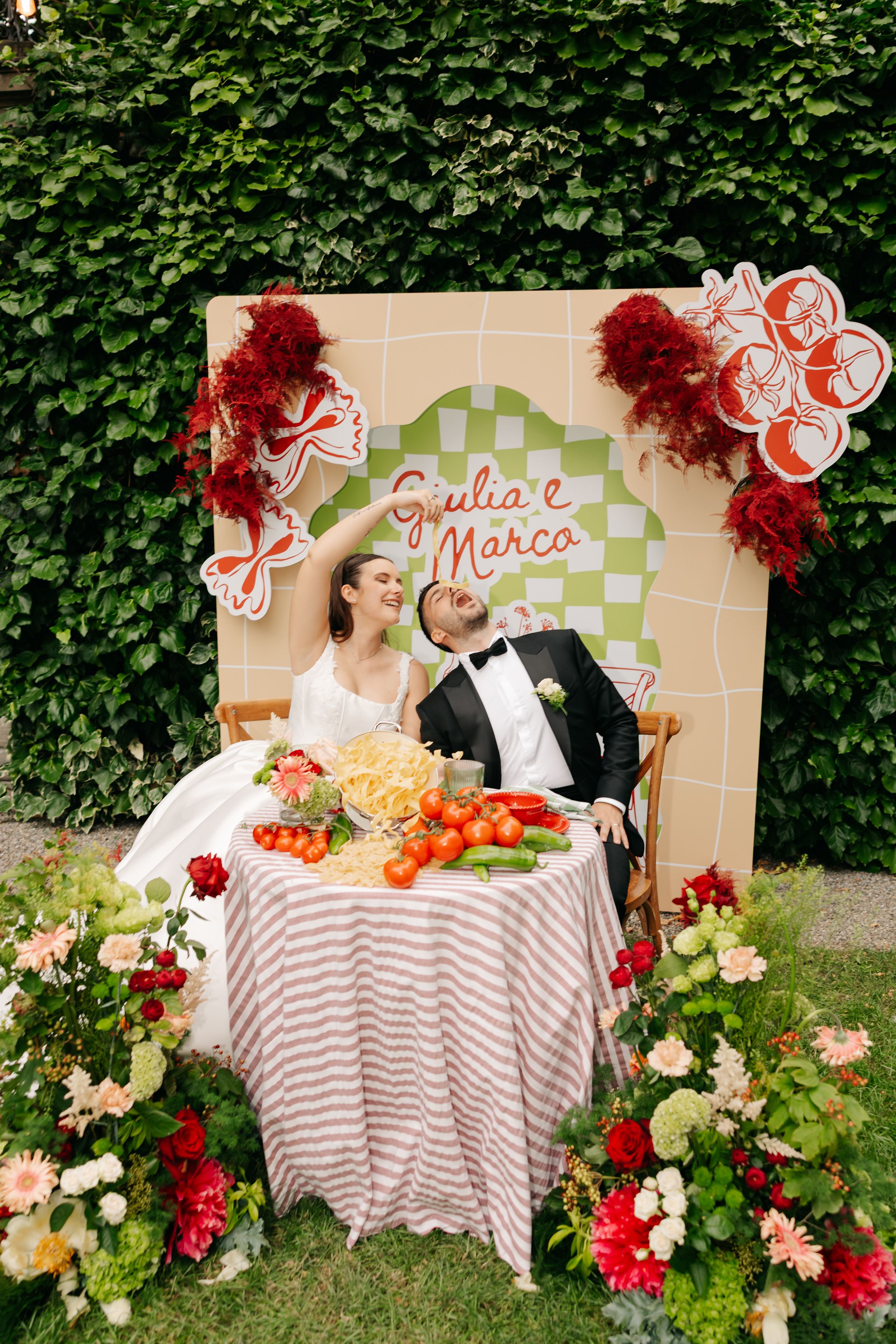 Italian themed wedding photo backdrop with table settings, red floral decorations and striped tablecloth against ivy wall.