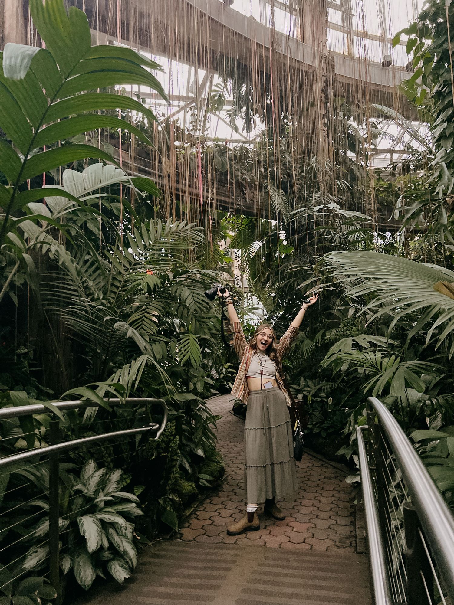 Person standing on a walkway surrounded by lush tropical plants in a greenhouse setting.