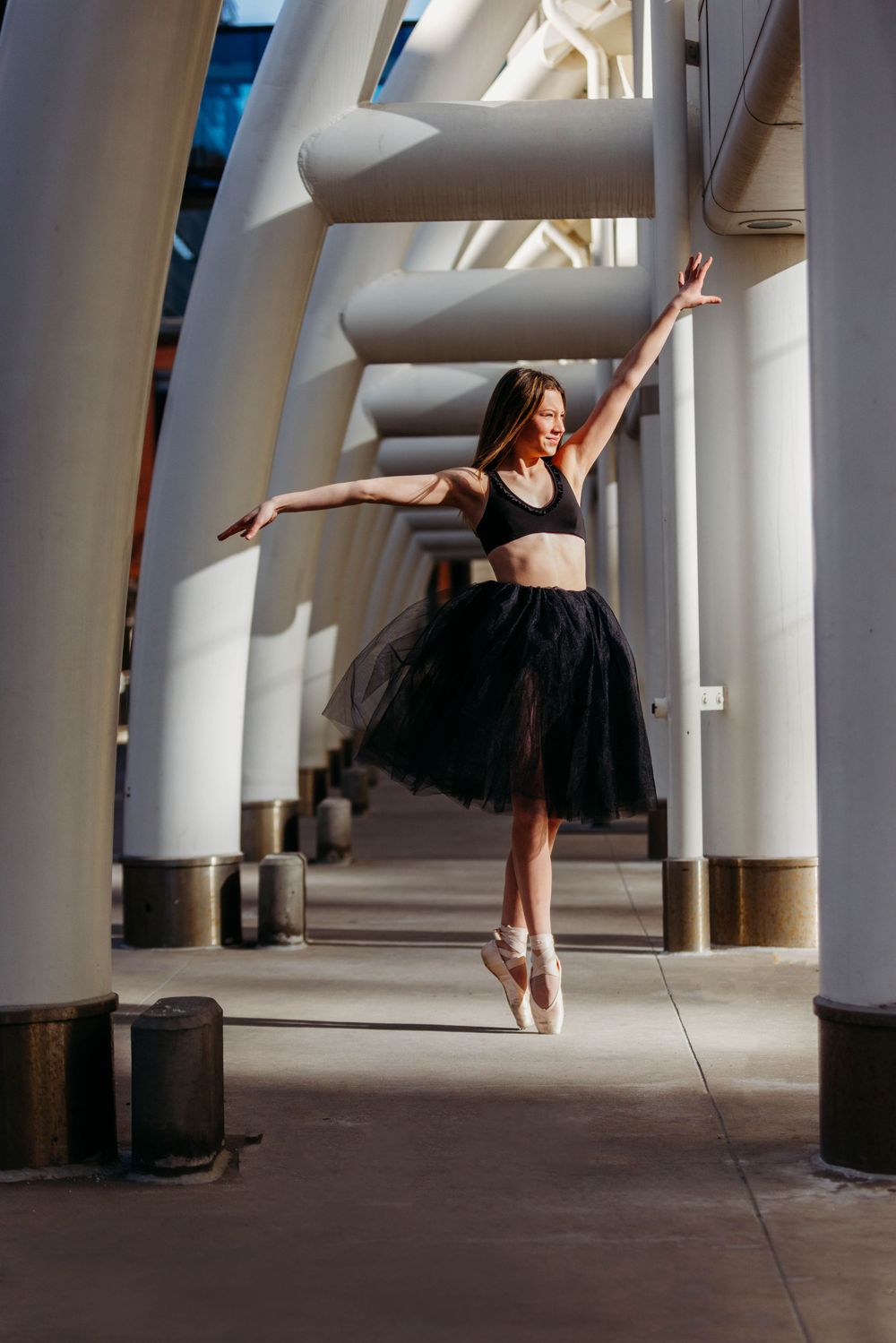 Ballet dancer in black tutu and pointe shoes performs graceful pose between architectural columns in natural light.
