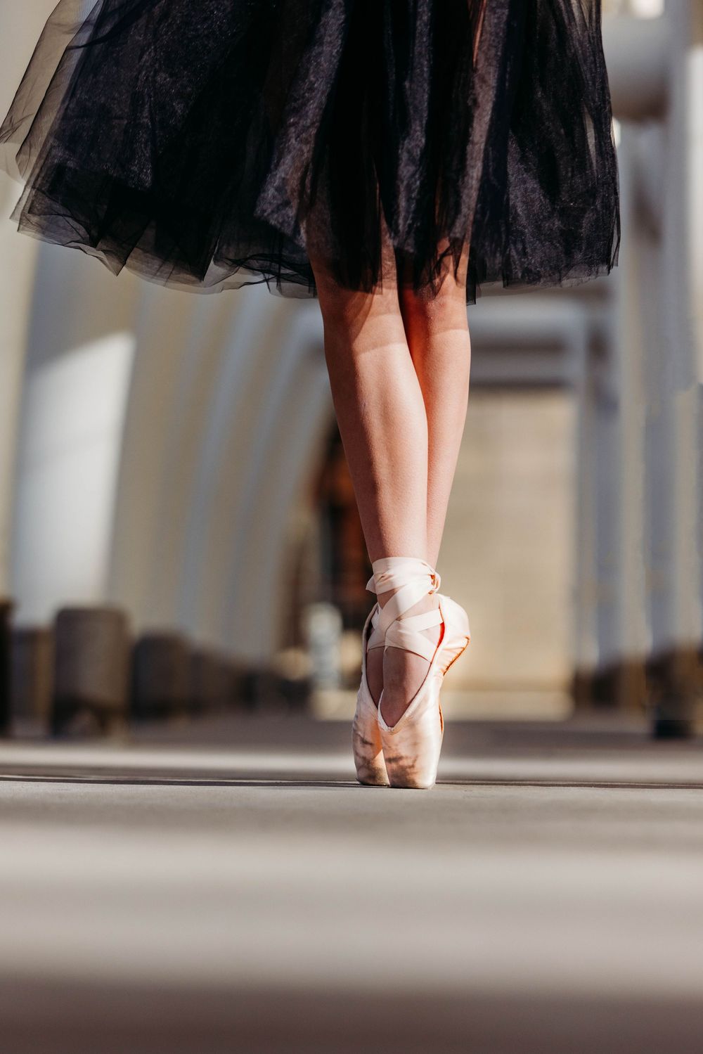 Ballet dancer in black tulle skirt stands en pointe in pink ballet shoes against blurred background.