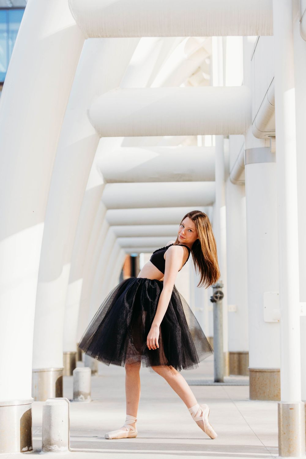 A dancer in a black tulle skirt poses gracefully in a white colonnade hallway with dramatic lighting and shadows.