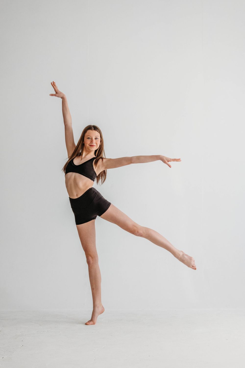 A dancer in black activewear performs a graceful high kick pose against a white studio backdrop.
