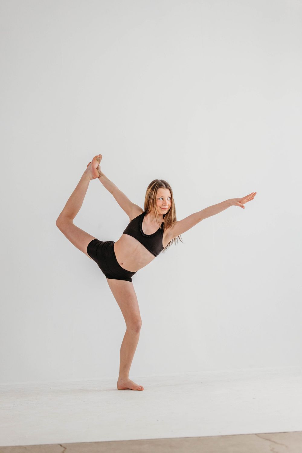 Dancer in black athletic wear performs a standing split pose against a white studio wall.