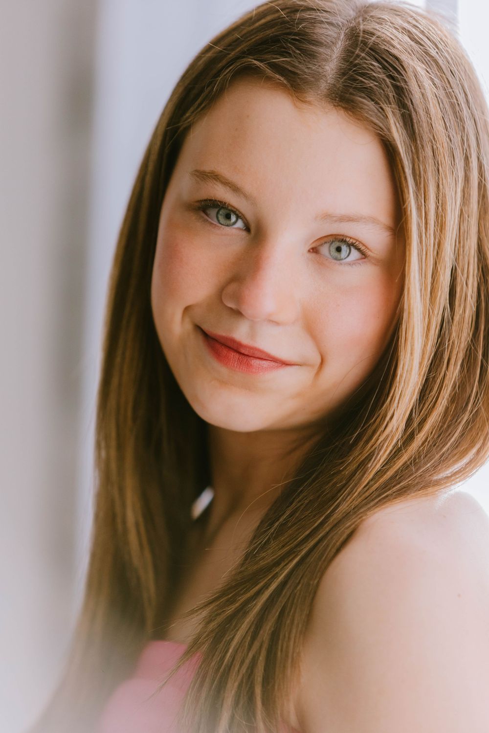 Portrait of a young person with long brown hair, light eyes, and a warm smile wearing a pink top.