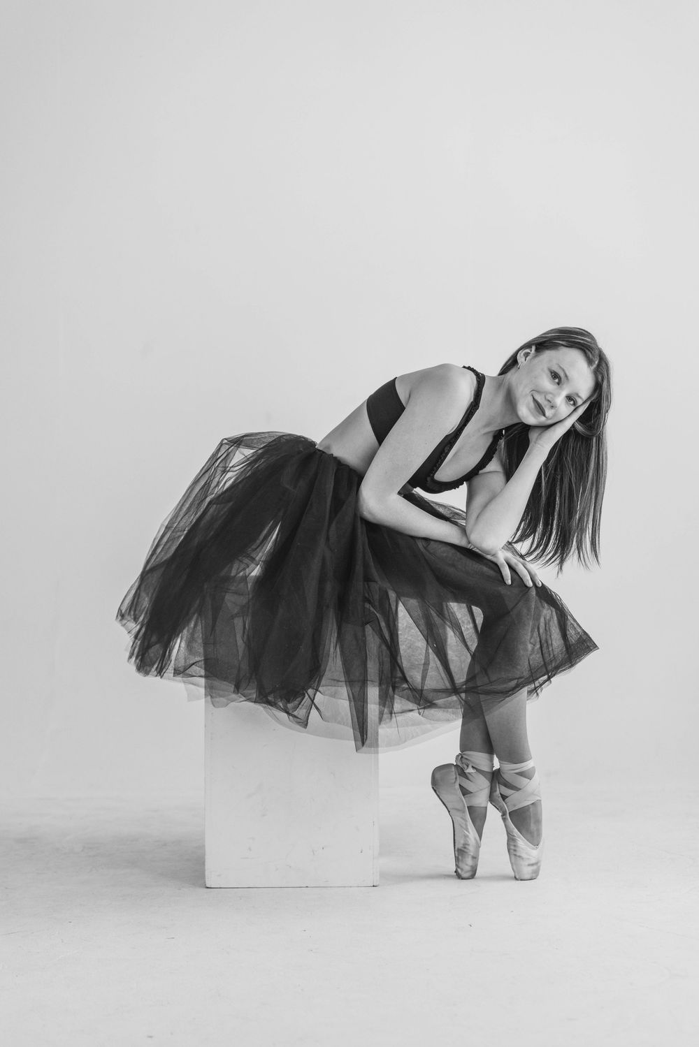 A ballerina in a black tulle skirt leans gracefully against a white cube in a minimalist studio setting.